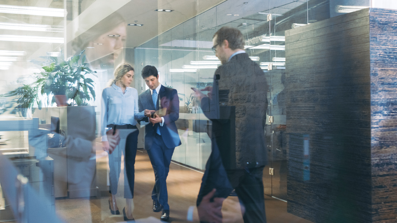 Businessman and Businesswoman Walking Through Glass Hallway, Discussing Work and Using Tablet Computer. Busy Corporate Office Building with Many Workers.